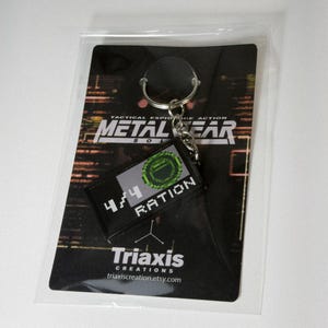 May include: A black keychain featuring a rectangular patch with the text "4/4 RATION" and a pixelated green design. The keychain is attached to a card with the words "METAL GEAR SOLID" and "Triaxis Creations" printed on it.