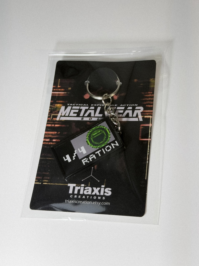May include: A black keychain featuring a rectangular patch with the text "4/4 RATION" and a pixelated green design. The keychain is attached to a card with the words "METAL GEAR SOLID" and "Triaxis Creations" printed on it.