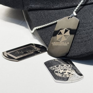Può includere: Tre targhette argentate con diversi design. Una ha il logo Umbrella Corporation, un'altra la scritta "CONDITION Fine", e la terza mostra "S.T.A.R.S. Raccoon Dep". Le targhette sono su una superficie bianca.
