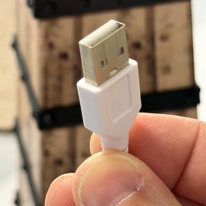 May include: A white USB cable with a silver-colored USB connector. The cable is held in a person's hand. The background is out of focus, but appears to be a wooden structure with black accents.