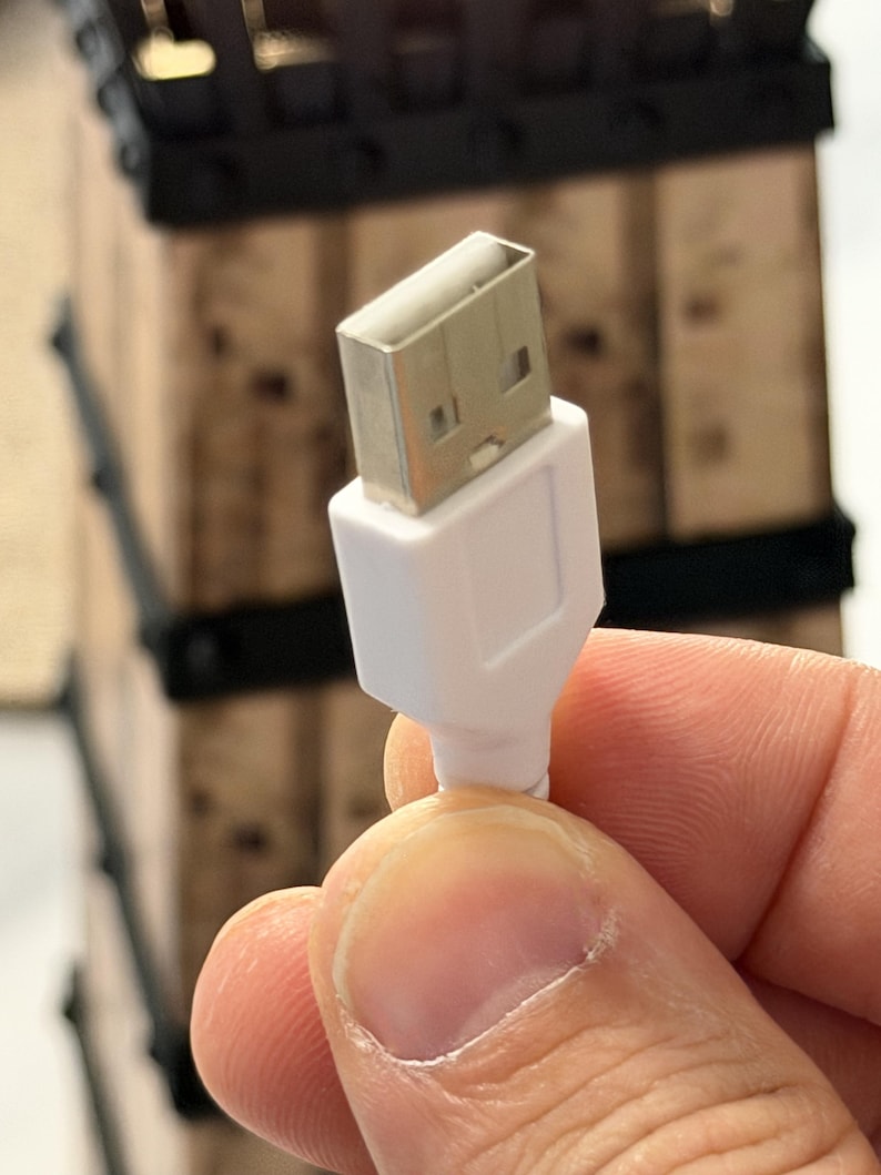 May include: A white USB cable with a silver-colored USB connector. The cable is held in a person's hand. The background is out of focus, but appears to be a wooden structure with black accents.