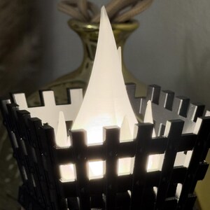 May include: A black and white decorative lantern with a bright white light inside. The lantern has a square shape with a fence-like design. A gold-colored vase with a rope handle is in the background.