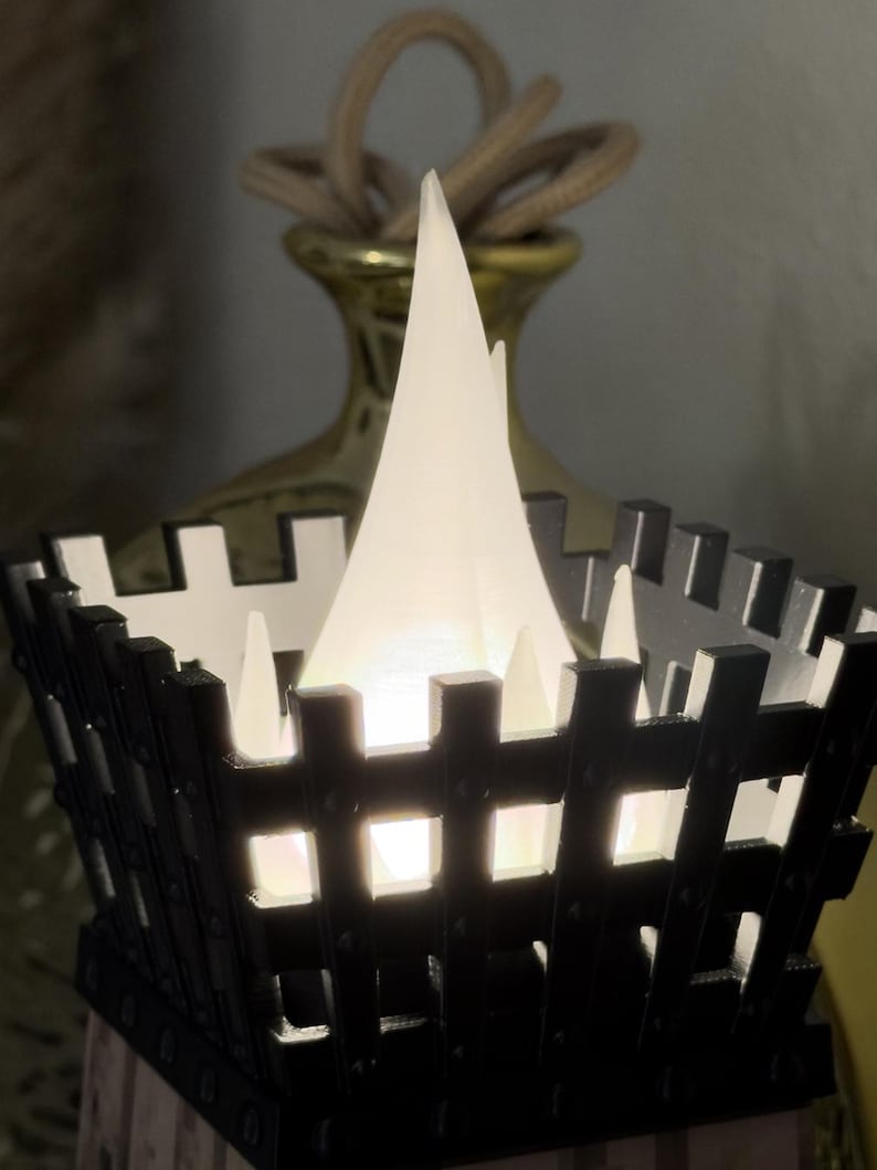 May include: A black and white decorative lantern with a bright white light inside. The lantern has a square shape with a fence-like design. A gold-colored vase with a rope handle is in the background.