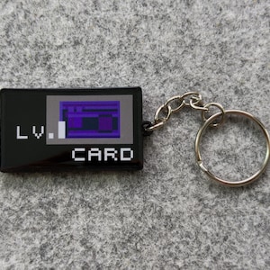 May include: Black rectangular keychain with the text "LV. CARD" in white. The card design features a purple and blue pixelated graphic. A silver chain and keyring are attached.