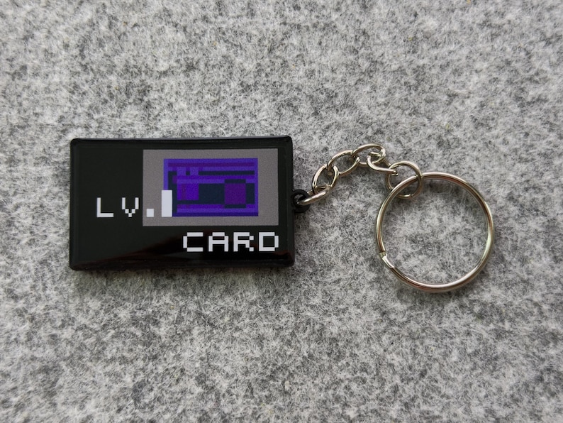 May include: Black rectangular keychain with the text "LV. CARD" in white. The card design features a purple and blue pixelated graphic. A silver chain and keyring are attached.