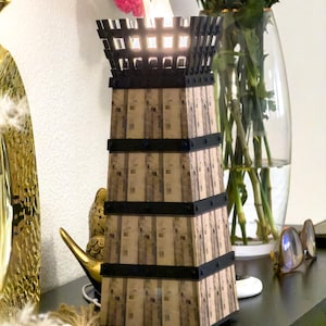 May include: A decorative tower-shaped lamp with a warm, inviting glow. The lamp features a textured, light brown exterior with black bands and a black top. The lamp is approximately 12 inches tall and is placed on a dark surface.