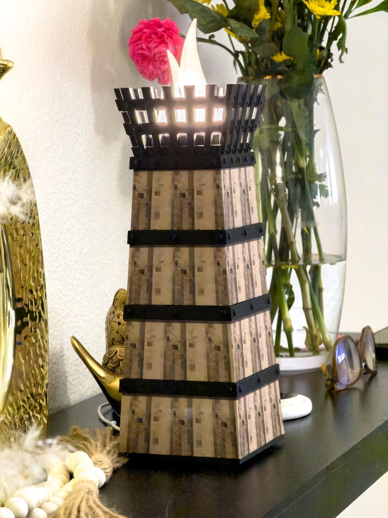 May include: A decorative tower-shaped lamp with a warm, inviting glow. The lamp features a textured, light brown exterior with black bands and a black top. The lamp is approximately 12 inches tall and is placed on a dark surface.