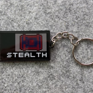 May include: Black rectangular keychain with the word "STEALTH" in white block letters. The keychain features a pixelated design of a red and blue object. A silver chain and keyring are attached.