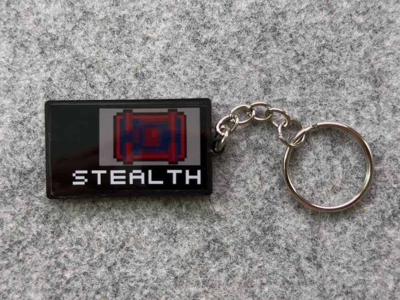 May include: Black rectangular keychain with the word "STEALTH" in white block letters. The keychain features a pixelated design of a red and blue object. A silver chain and keyring are attached.