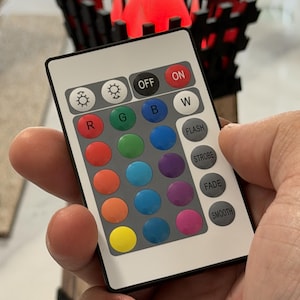 May include: A white remote control with multiple color buttons is held in a hand. The remote has buttons labeled "ON", "OFF", "W", "R", "G", "B", and others for color selection and lighting effects. A decorative wooden structure with a red light is in the background.