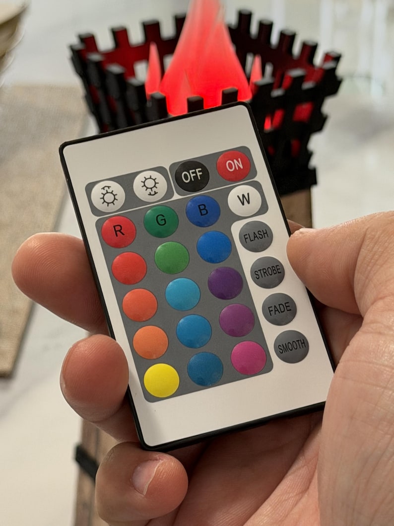 May include: A white remote control with multiple color buttons is held in a hand. The remote has buttons labeled "ON", "OFF", "W", "R", "G", "B", and others for color selection and lighting effects. A decorative wooden structure with a red light is in the background.