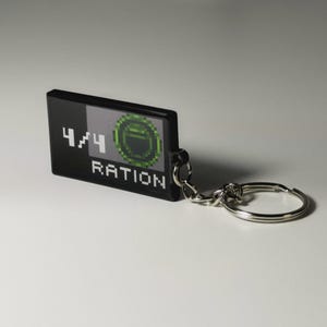 May include: Black rectangular keychain with the text "4/4 RATION" in white. The keychain features a pixelated green and gray design. A silver key ring and chain are attached.