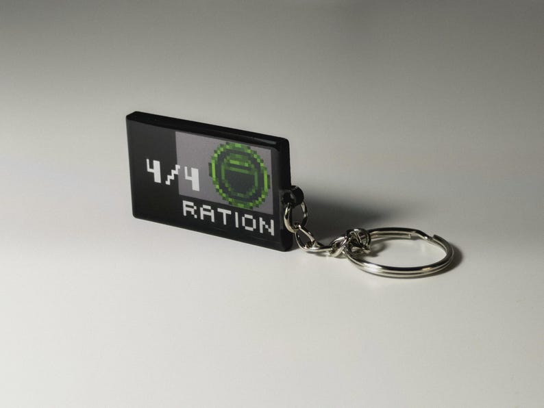 May include: Black rectangular keychain with the text "4/4 RATION" in white. The keychain features a pixelated green and gray design. A silver key ring and chain are attached.