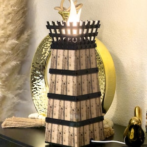 May include: A decorative lamp with a tower design. The lamp has a light brown, textured body with black bands and a black top. The top has a flame-like light. The lamp is on a black surface.