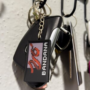 May include: A keychain with a black rectangular tag that reads "BANDANA" in white capital letters. The tag features a pixelated red design. The keychain is attached to a car key and other keys.