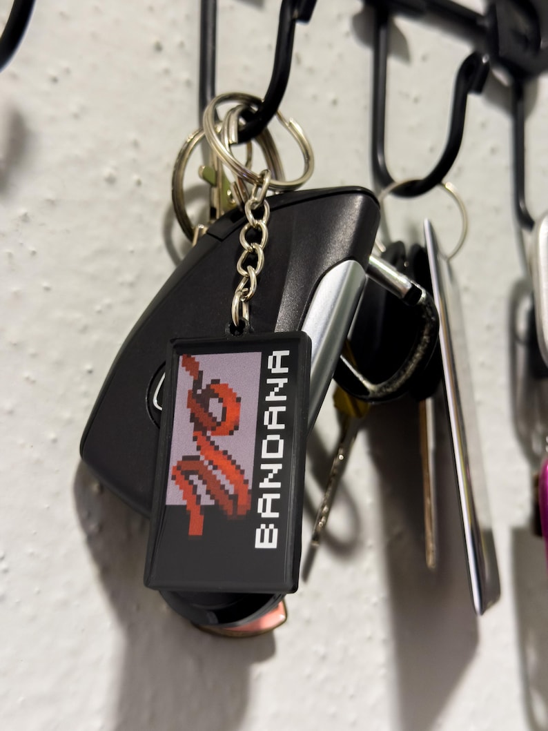 May include: A keychain with a black rectangular tag that reads "BANDANA" in white capital letters. The tag features a pixelated red design. The keychain is attached to a car key and other keys.