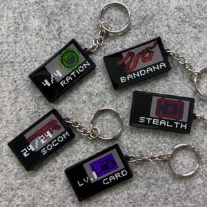 May include: A collection of black keychains with pixelated graphics and text. Each keychain features a different design, including "RATION," "BANDANA," "STEALTH," "SOCOM," and "LV. 1 CARD." The keychains have silver key rings and chains.