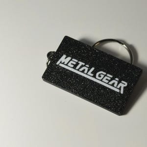 May include: A black rectangular keychain with the words "METAL GEAR" in white, block letters. The keychain has a textured surface and a silver key ring attached.