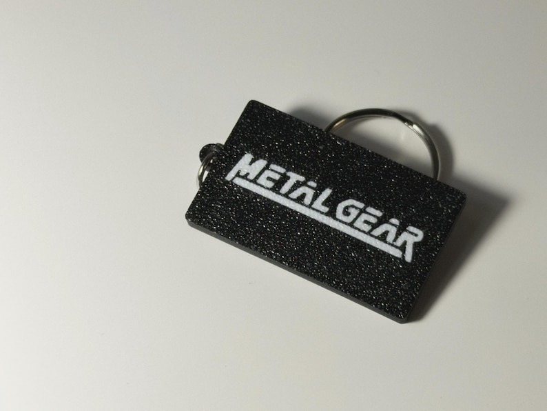 May include: A black rectangular keychain with the words "METAL GEAR" in white, block letters. The keychain has a textured surface and a silver key ring attached.