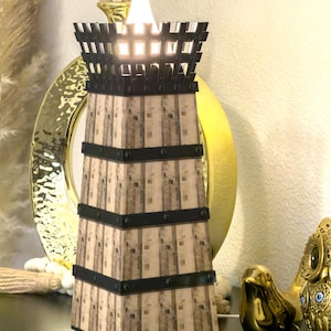 May include: A decorative lamp with a tower design. The lamp has a light brown, textured body with black bands and a black top. A bright white light shines from the top. The lamp is on a black surface with other decorative items.