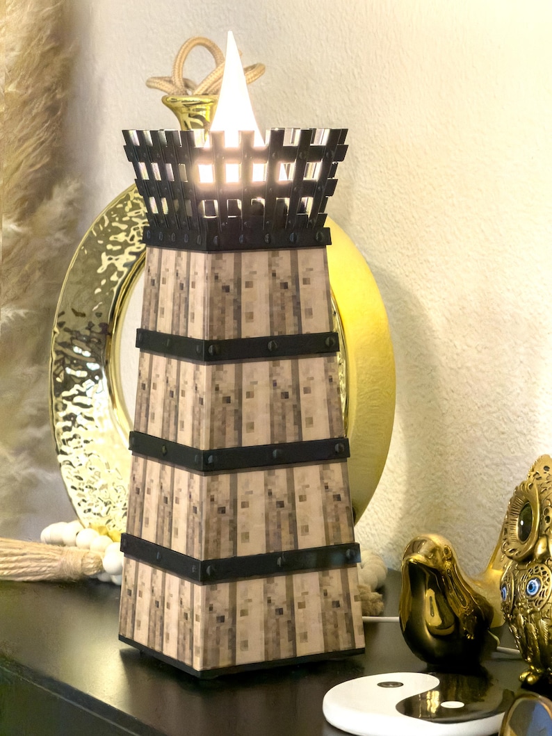 May include: A decorative lamp with a tower design. The lamp has a light brown, textured body with black bands and a black top. A bright white light shines from the top. The lamp is on a black surface with other decorative items.