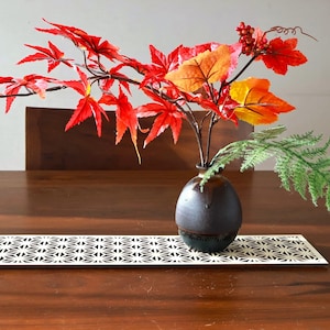 Puede incluir: Un arreglo decorativo con vibrantes hojas de arce rojas y naranjas, frondas de helecho verdes y pequeñas bayas rojas en un jarrón de cerámica marrón oscuro. El jarrón está sobre una mesa de madera con un camino de mesa estampado en blanco y negro.