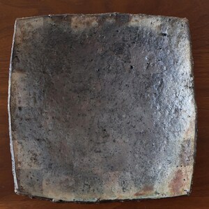 Puede incluir: Plato cuadrado de cerámica con una superficie texturizada y moteada en tonos grises y marrones. Los bordes tienen un color ligeramente más claro, y el diseño general es rústico y hecho a mano.
