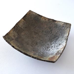 May include: A square, handmade ceramic dish with a slightly curved shape. The dish has a textured, mottled surface with shades of gray, brown, and hints of silver. The edges are rough and uneven, giving it a rustic appearance.