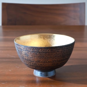 May include: A ceramic bowl with a gold interior and a textured, dark brown exterior. The bowl has a small, dark base and sits on a wooden surface. The bowl's interior reflects light, creating a warm glow.