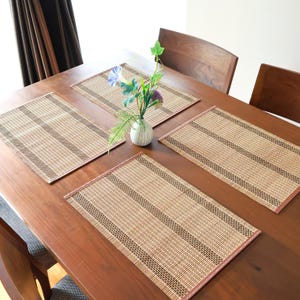 May include: Four rectangular bamboo placemats are arranged on a wooden table. Each mat has a light brown colour with darker brown stripes and a reddish-brown border. A small vase with flowers sits in the centre.