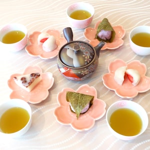 Può includere: Un set da tè giapponese con una teiera floreale, piatti rosa a forma di fiore e piccole tazze bianche. I piatti contengono mochi e altri dolci tradizionali. Il tè è di colore verde chiaro.