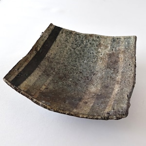 May include: A square ceramic dish with a curved, concave shape. The surface features a mottled, textured finish in shades of gray, brown, and muted green, with a distinct dark brown vertical stripe running along one side of the piece.
