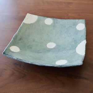 May include: A square ceramic plate with a light blue-green glaze and large, irregular white polka dots. The plate has a slightly uneven edge, giving it a handmade appearance. The plate is on a wooden surface.