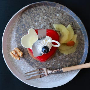 May include: A heart-shaped dessert with a glossy red glaze, topped with white cream, a blueberry, and a white chocolate heart. Served on a speckled plate with kiwi slices, an almond, and a walnut. A fork with a wooden handle is also present.