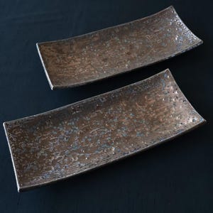 Könnte beinhalten: Zwei rechteckige Serviertabletts in Bronzefarbe mit strukturierter Oberfläche und leicht gebogenen Kanten. Die Tabletts haben einen metallischen Glanz und werden auf dunklem Hintergrund präsentiert. Geeignet zum Servieren von Vorspeisen oder kleinen Gerichten.