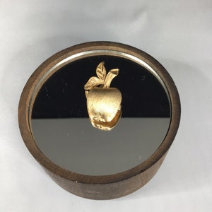 K&ouml;nnte beinhalten: Eine goldfarbene Apfelnadel mit einem Blatt liegt auf einer verspiegelten Oberfl&auml;che in einer Holzkiste.