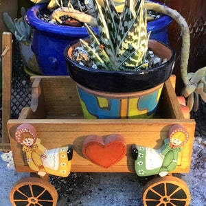 May include: A wooden wagon planter with two painted figures on the front. The wagon is painted in shades of brown, yellow, and green. The figures are wearing red, yellow, and green clothing. The wagon is filled with a potted succulent plant.