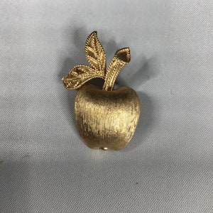 K&ouml;nnte beinhalten: Eine goldfarbene Apfelbrosche mit strukturierter Oberfl&auml;che und einem Stiel mit zwei Bl&auml;ttern.