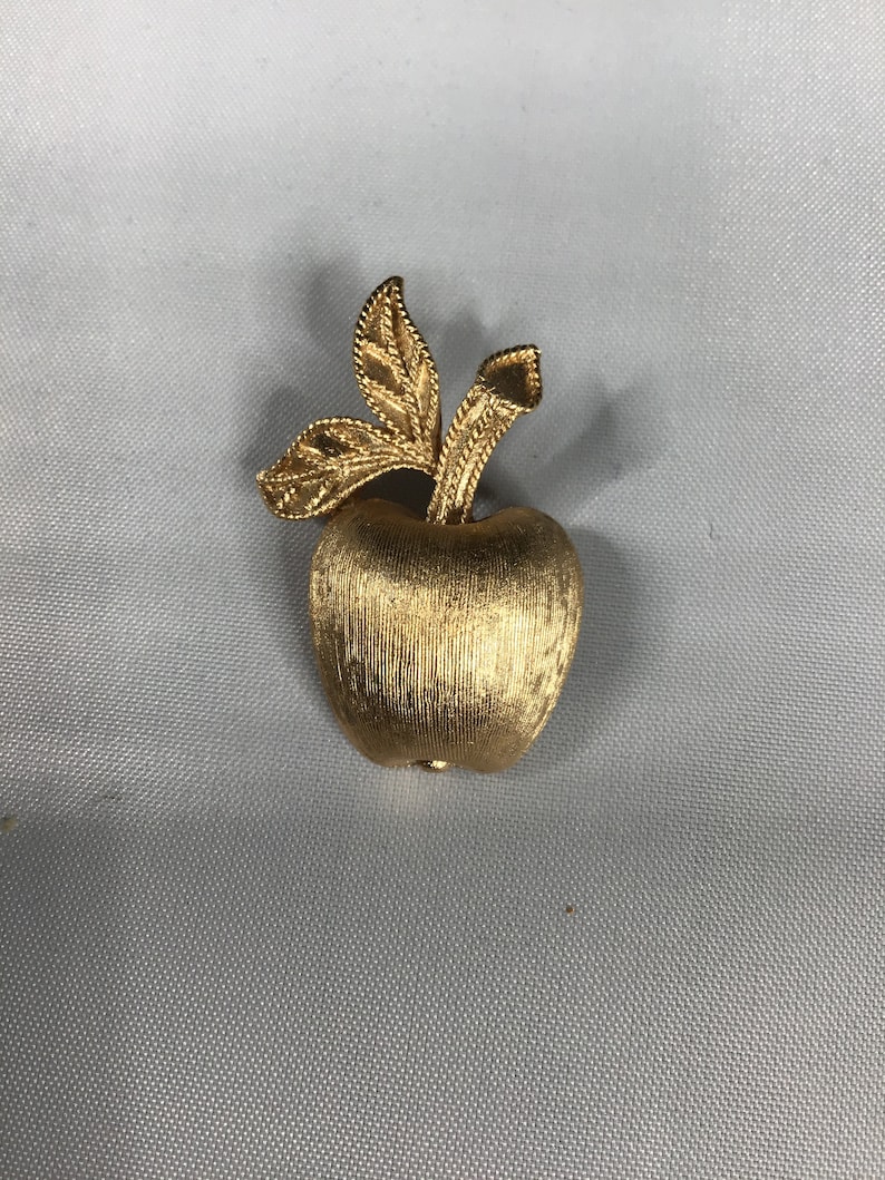 K&ouml;nnte beinhalten: Eine goldfarbene Apfelbrosche mit strukturierter Oberfl&auml;che und einem Stiel mit zwei Bl&auml;ttern.