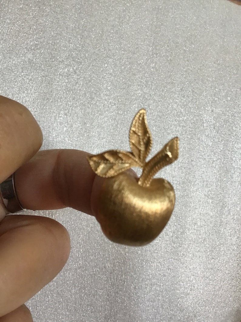 K&ouml;nnte beinhalten: Eine goldfarbene Apfelbrosche mit einem detaillierten Blattdesign. Die Brosche ist ein schlichtes und elegantes Schmuckst&uuml;ck.