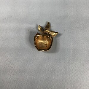 K&ouml;nnte beinhalten: Goldfarbene Apfelbrosche mit Stiel und Bl&auml;ttern. Die R&uuml;ckseite der Brosche hat ein kleines, rundes, goldfarbenes Etikett mit dem Wort "Avon" darauf.