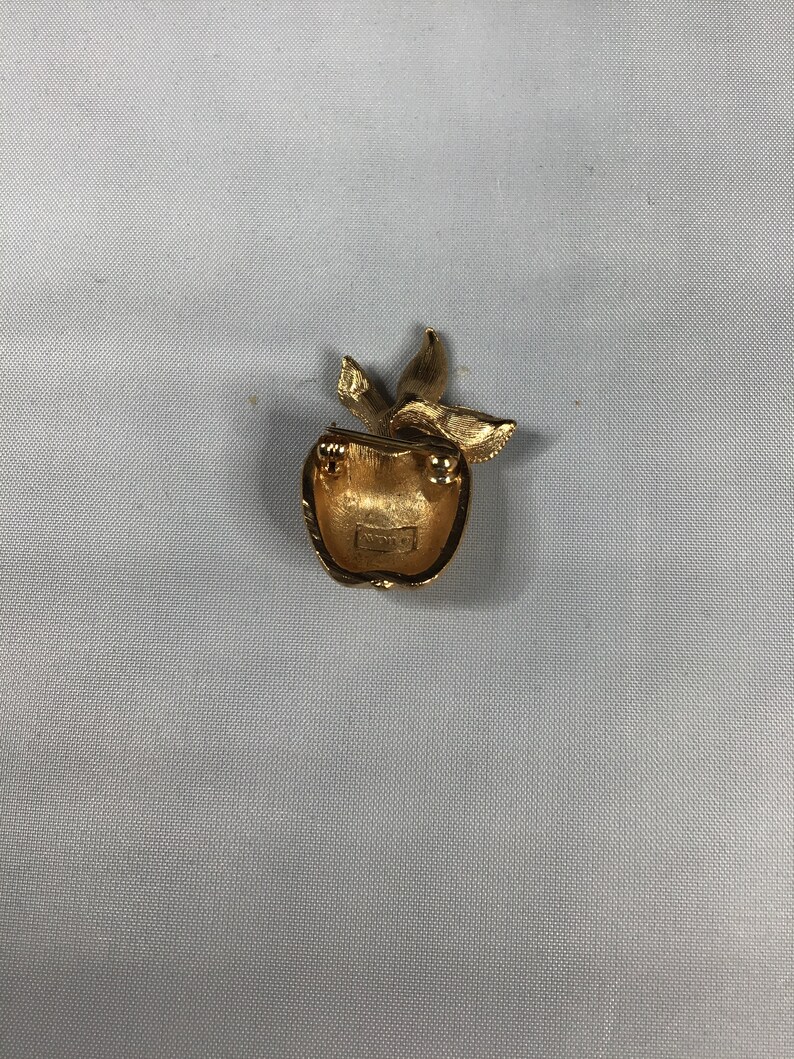 K&ouml;nnte beinhalten: Goldfarbene Apfelbrosche mit Stiel und Bl&auml;ttern. Die R&uuml;ckseite der Brosche hat ein kleines, rundes, goldfarbenes Etikett mit dem Wort "Avon" darauf.