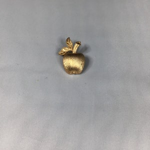 K&ouml;nnte beinhalten: Eine goldfarbene Apfelbrosche mit strukturierter Oberfl&auml;che und einem einfachen Blattdesign.