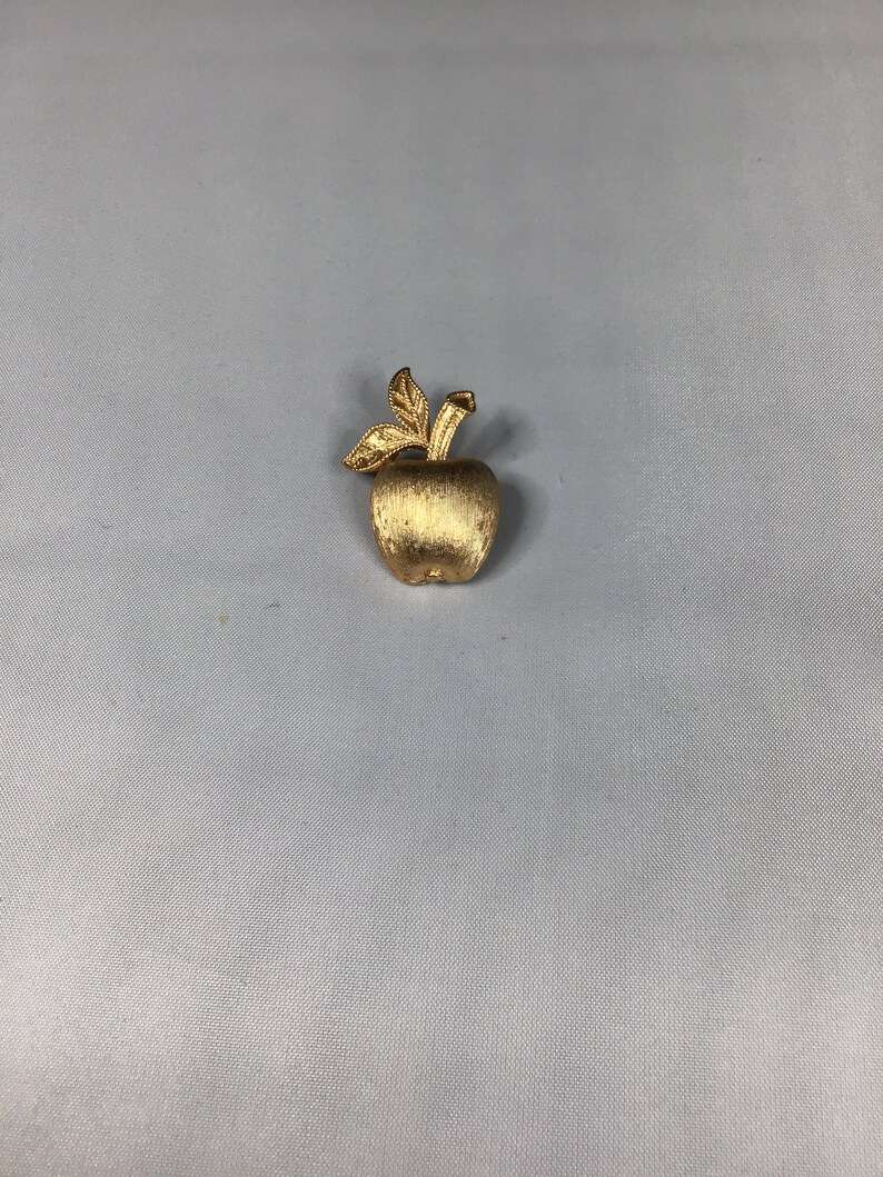 K&ouml;nnte beinhalten: Eine goldfarbene Apfelbrosche mit strukturierter Oberfl&auml;che und einem einfachen Blattdesign.