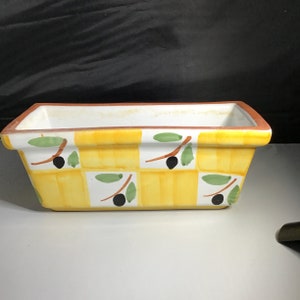 Puede incluir: Una maceta de cerámica amarilla y blanca con un patrón a cuadros y diseños de ramas de olivo. La maceta tiene una forma rectangular y es perfecta para exhibir plantas o flores.