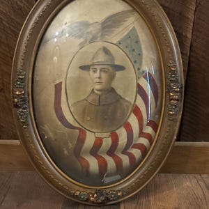May include: An oval-shaped, ornate gold-toned frame containing a sepia-toned portrait of a person wearing a military-style hat and uniform. Behind the portrait, a large American flag and an eagle are depicted. A small white tag with the number 6009 is visible.