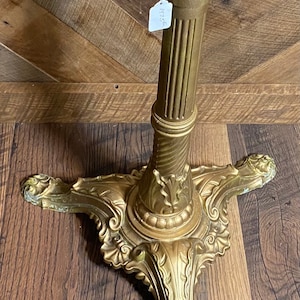May include: Ornate gold-colored table base with a round top and decorative, scrolling base. The base has a fluted column and detailed floral and leaf patterns. The top is painted yellow with visible screw holes.