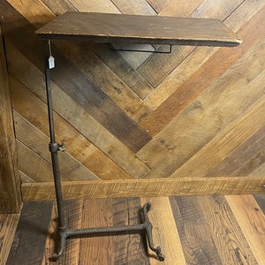 May include: An antique adjustable table with a dark wooden top and a metal base. The table's height is adjustable, and it has a sturdy, ornate base. The wooden top is rectangular and shows signs of wear.