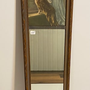 May include: A vintage wooden framed mirror with a carved design. The mirror reflects a white wall with vertical wood paneling. The top half of the mirror shows a framed print of a woman and child standing by a window with a dog. The print is in sepia tones.