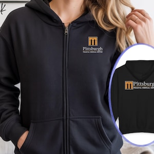 May include: Black zip-up hoodie with a hood and front pockets. The hoodie features the text "Pittsburgh Trauma Medical Center" and a logo on the chest. The word "Black" is written in the upper left corner.